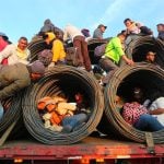 Migrants ride on a truckload of steel rolls.