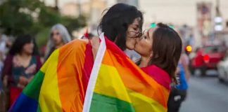 Two women kissing in the street wrapped in a multicolored rainbow gay pride flag.