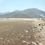 The dust and mud of a dry resevoir with a small amount of water visible in the distance. Even farther away in the background dry, rocky mountains are visible.