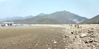 The dust and mud of a dry resevoir with a small amount of water visible in the distance. Even farther away in the background dry, rocky mountains are visible.