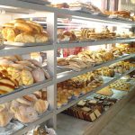 Shelves of a Mexican bakery, filled with different kinds of sweet bread. Behind, people look in the window.