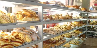 Shelves of a Mexican bakery, filled with different kinds of sweet bread. Behind, people look in the window.