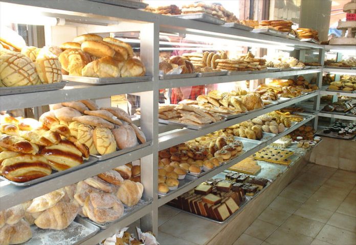Shelves of a Mexican bakery, filled with different kinds of sweet bread. Behind, people look in the window.