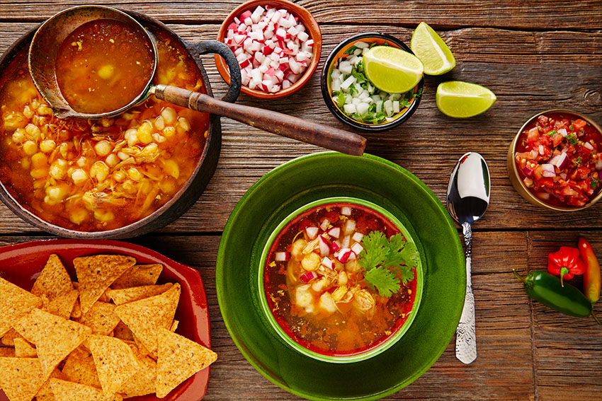 A bowl of pozole surrounded by onion, lime and other condiments.