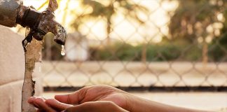 Hands cupped under a dripping tap, outdoors with palm trees and a fence in the background