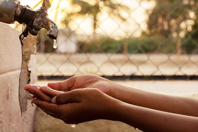 Hands cupped under a dripping tap, outdoors with palm trees and a fence in the background