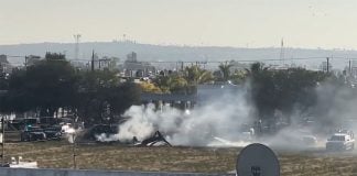 Burning rubbish from the helicopter crash sits in a field surrounded by police cars. The tail of the helicopter is visible between plumes of smoke.