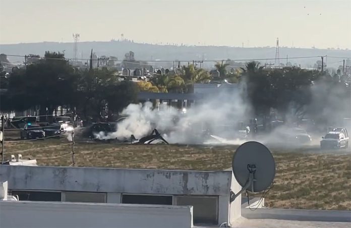 Burning rubbish from the helicopter crash sits in a field surrounded by police cars. The tail of the helicopter is visible between plumes of smoke.