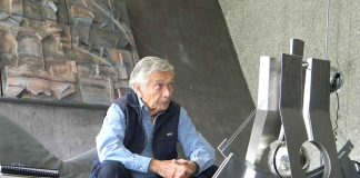 Agustín Hernández sits on a couch in a grey room, behind a coffee table with an architectural model.