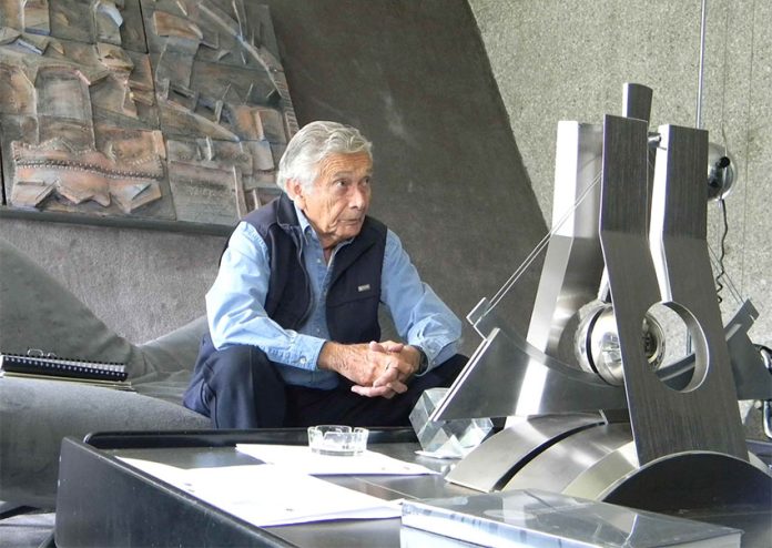Agustín Hernández sits on a couch in a grey room, behind a coffee table with an architectural model.