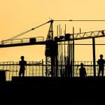 The silhouette of construction workers and a crane against a yellow sky.