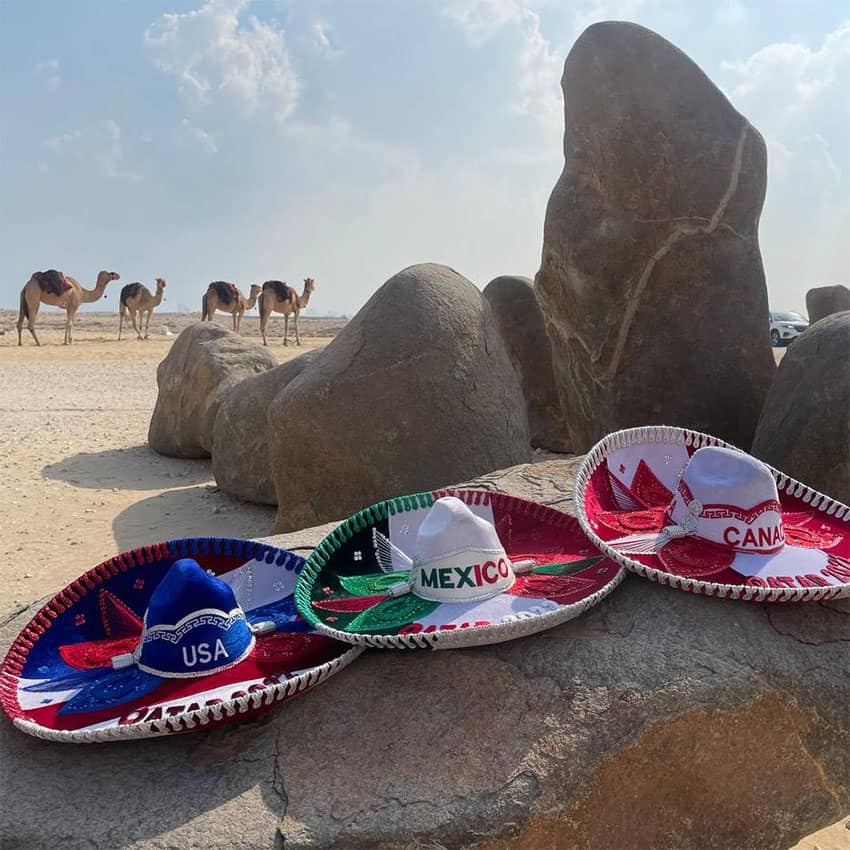 Three mariachi hats decorated with the colors of each 2026 host country, in the desert of Qatar with camels and rocks in the background.