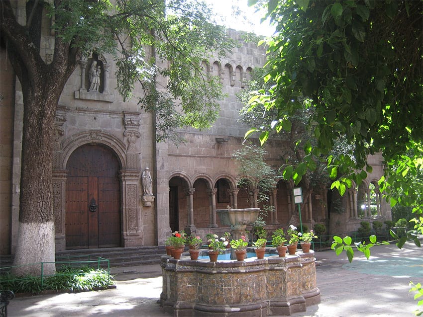 An old stone building surrounded by a fountain and lush gardens.