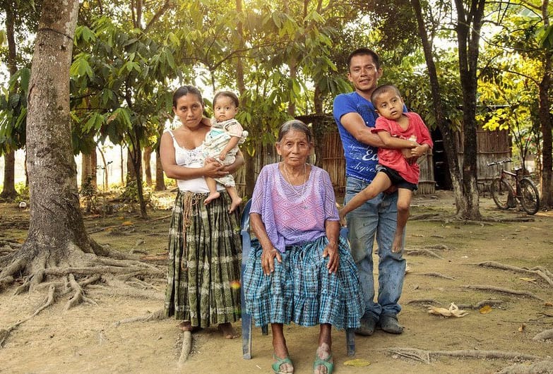 An indigenous Maya family poses in an outdoor setting. The mother and father stand, each holding a child, whle the grandmother remains seated.