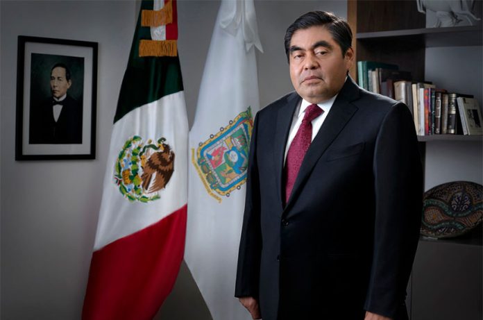 Miguel Barbosa Huerta in his office with a Mexican flag, a Puebla flag and a portrait of Benito Juárez.
