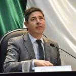 Pablo Monroy sits at a desk with a microphone, wearing a gray suit.