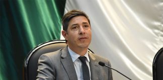 Pablo Monroy sits at a desk with a microphone, wearing a gray suit.