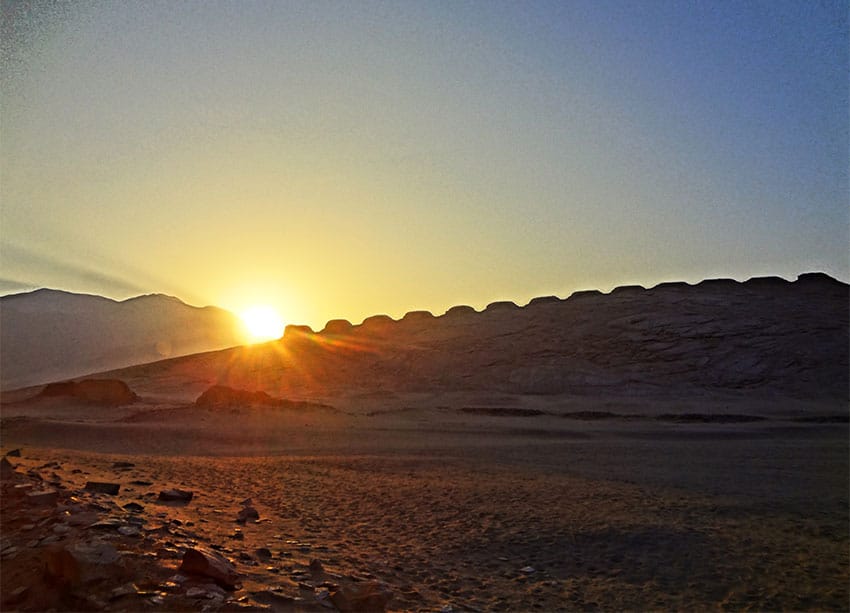 The sun peaks over a ridge that has a line of short towers built along it, in the desert.