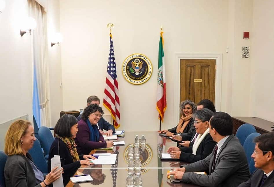 The Mexican and U.S. delegations sit at a conference table with U.S. and Mexican flags in the background.