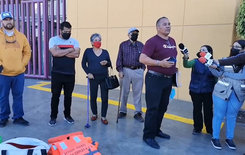 A small group of men an women stand in front of a pile of life vests and whale photos in front of a building, while a man in a red shirt speaks into the microphones of several news reporters.