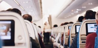 A well-lit aisle inside an airplane with people seated, seen from the back of the plane.
