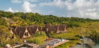 The modular rooms of Habitas Bacalar, made of what appears to be palm thatch, look out over a river or estuary with forest in the background.