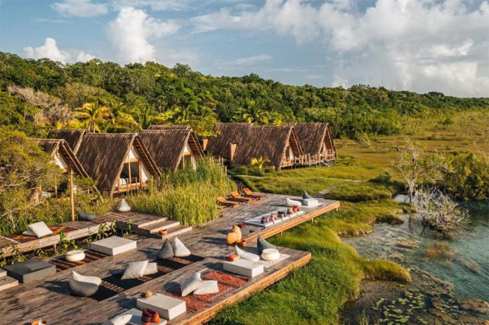 The modular rooms of Habitas Bacalar, made of what appears to be palm thatch, look out over a river or estuary with forest in the background.
