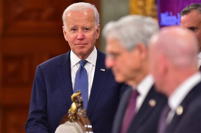 Biden in a wood paneled room, with two out-of-focus officials in the foreground