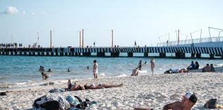 A dozen or so people lie and stand on the beach with clear blue water and a long pier in the background.