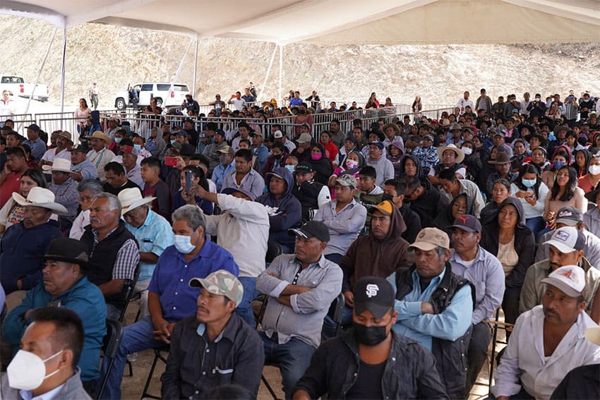 In a white shade tent with rocks visible outside, a crowd of people with serious expressions look toward the camera.