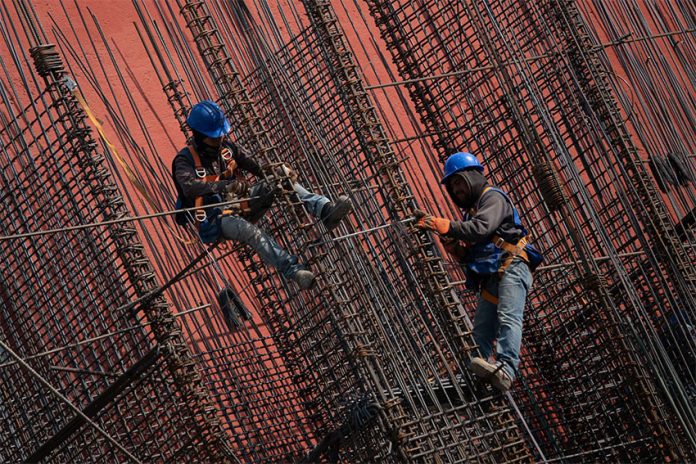 Two construction workers working suspended among steel beams on an orange background.