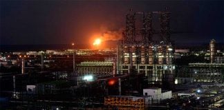 A gas flare burns at night in the middle of a large industrial complex, with the dark sea barely visible in the background.