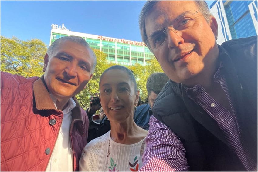 A selfie of the three politicians outdoors with trees and a multistory building in the background.