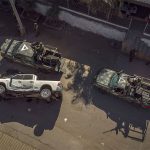 An aerial view of two camo vehicles driving past a flipped white pickup truck with bullet holes on a dirt road, in front of a house.