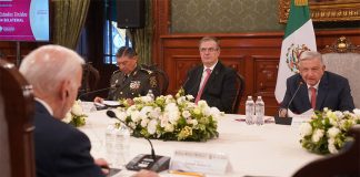 López Obrador speaks into a microphone while seated at a table with officials seated at his side and flowers in the center.