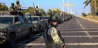A soldier in uniform looks at the camera, while dozens of army vehicles carrying soldiers line the highway in the background.