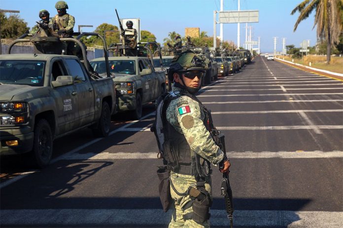 A soldier in uniform looks at the camera, while dozens of army vehicles carrying soldiers line the highway in the background.