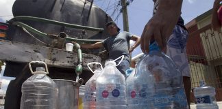 In the background, a man in a baseball cap fills a large pot from a tube coming out of a water truck. In the foreground, dozens of empty 5 or 10-liter plastic bottles wait to be filled.