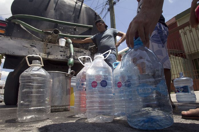 In the background, a man in a baseball cap fills a large pot from a tube coming out of a water truck. In the foreground, dozens of empty 5 or 10-liter plastic bottles wait to be filled.
