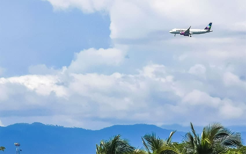 A Volaris airplane flies over palm trees with mountains in the background on a partly cloudy day.