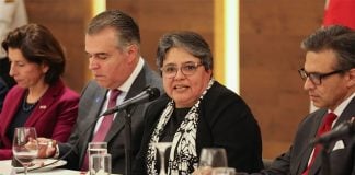 A woman speaks into a microphone while seated at a table next to other formally dressed politicians.