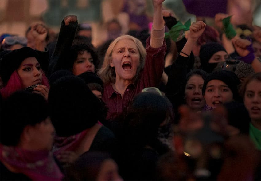 An older light-skinned women, yelling, reaches out from the center a crowd of other women.