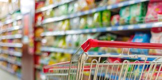 A shopping cart in the foreground with a colorful, out-of-focus grocery isle in the background.