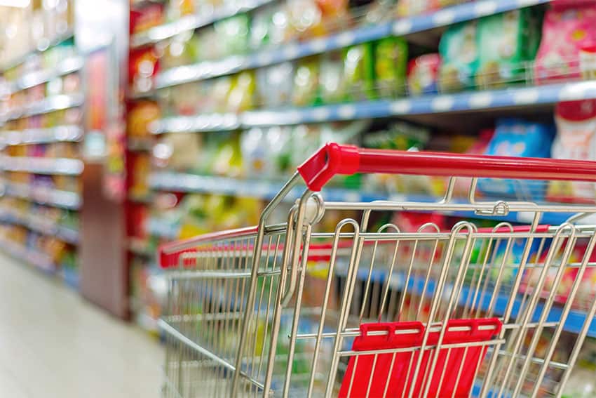 A shopping cart in the foreground with a colorful, out-of-focus grocery isle in the background.