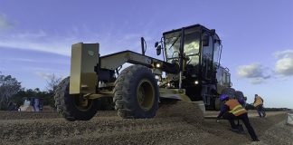 Construction workers use heavy machinery to smooth gravel, with forest in the background.