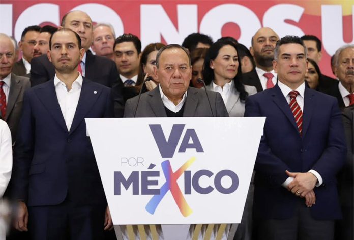 Politicians in suits with frowns and serious expressions stand behind a podium with the Va por México logo.