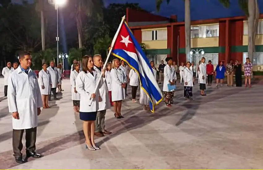 Cuban doctors standing in rows, one holding a Cuban flag, in a government sendoff ceremony to send the doctors to work in Mexico