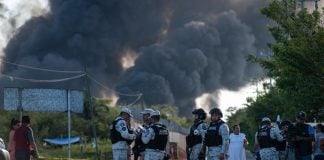 National Guard members in uniform stand on a cordoned-off road in a wooded area with billowing smoke blocking most of the sky in the background.