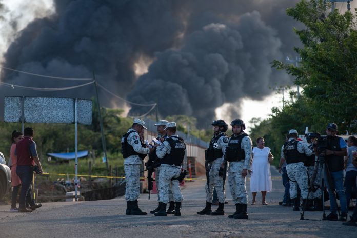 National Guard members in uniform stand on a cordoned-off road in a wooded area with billowing smoke blocking most of the sky in the background.
