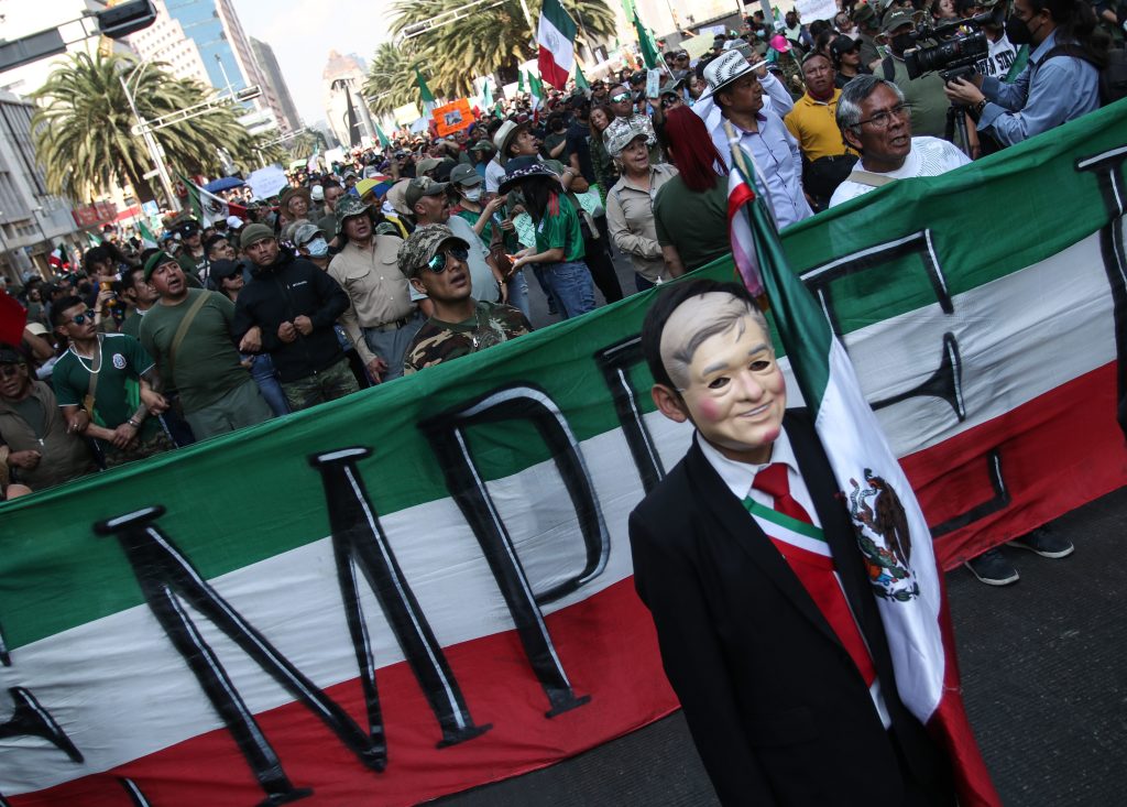 A protester dressed satirically in a mask and suit as President López Obrador stands in front of a tricolor banner with a crowd waving Mexican flags behind. In the distance is the Mexico City Angel of Independence landmark.
